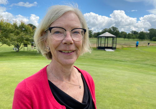 Blond kvinna i övre medelåldern, med röd tröja  och glasögon, står utomhus på en golfbana. 